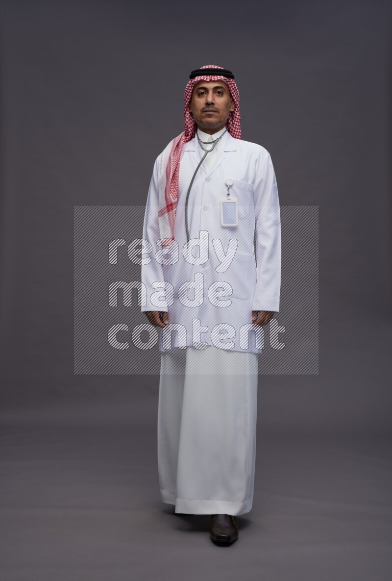 Saudi man wearing thob with lab coat and shomag with pocket employee badge with stethoscope standing interacting with the camera on gray background