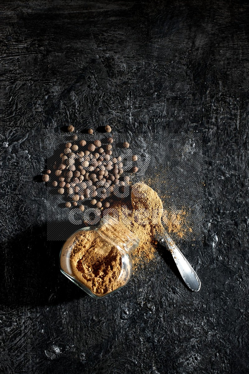 A flipped glass spice jar and metal spoon full of allspice powder and powder spilled out of it with whole balls on a textured black flooring