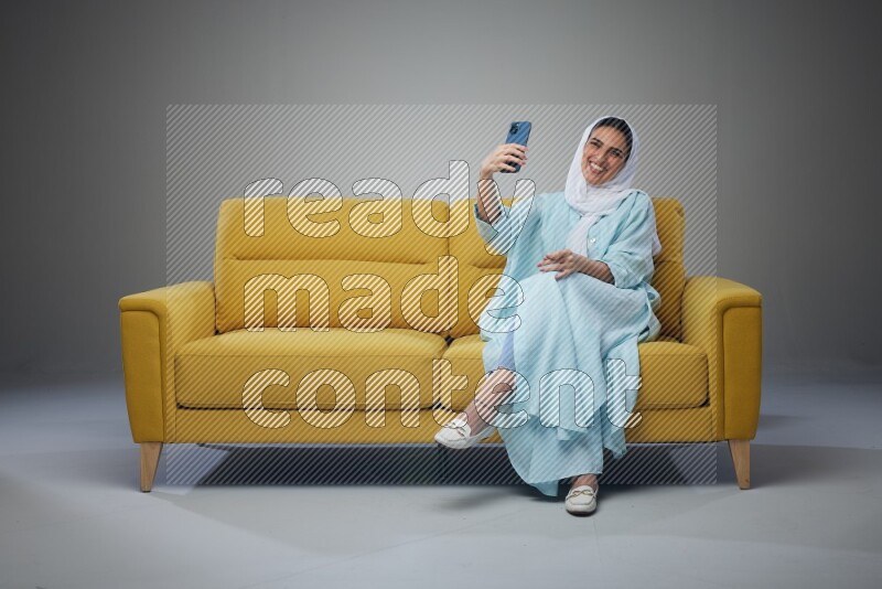 A Saudi woman wearing a light blue Abaya and white head scarf sitting on a yellow sofa and taking selfies with her phone eye level on a grey background