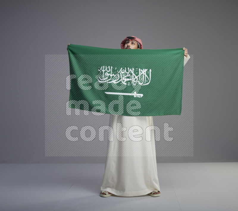 A saudi man standing wearing thob and red shomag holding big saudi flag on gray background