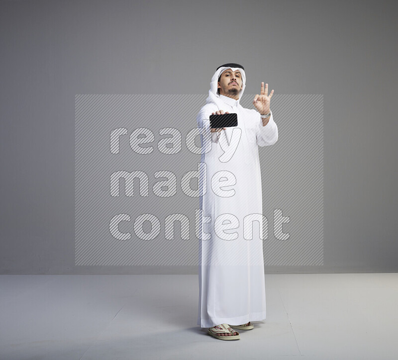 A Saudi man standing wearing thob and white shomag showing phone to camera on gray background