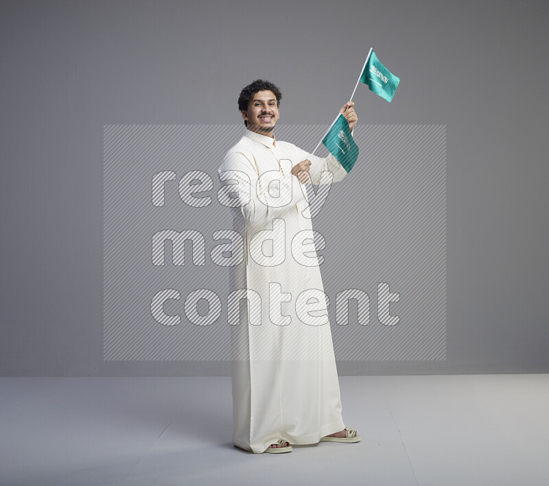A Saudi man standing wearing thob holding small Saudi flag on gray background