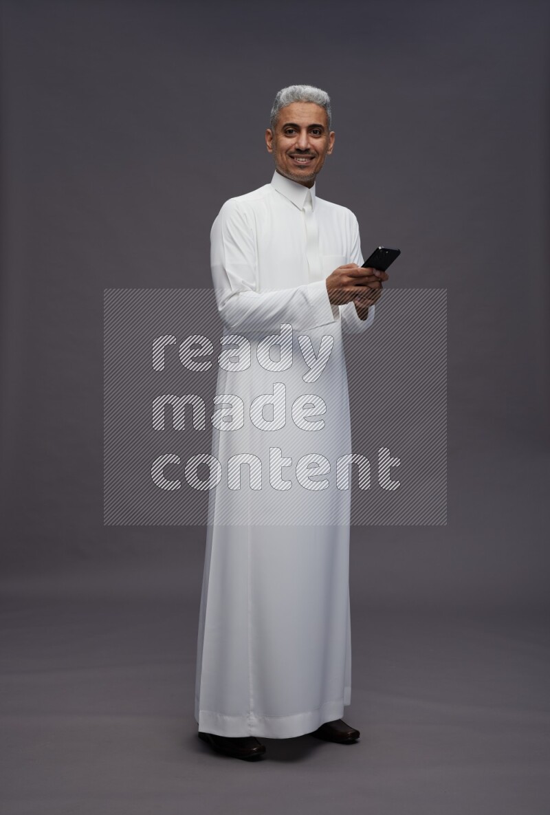 Saudi man wearing thob standing texting on phone on gray background