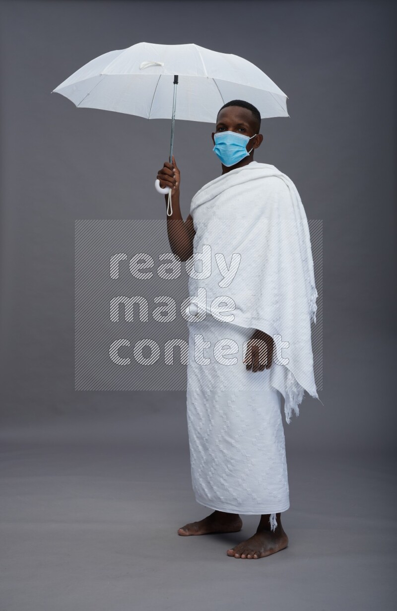 A man wearing Ehram with face mask Standing holding umbrella on gray background