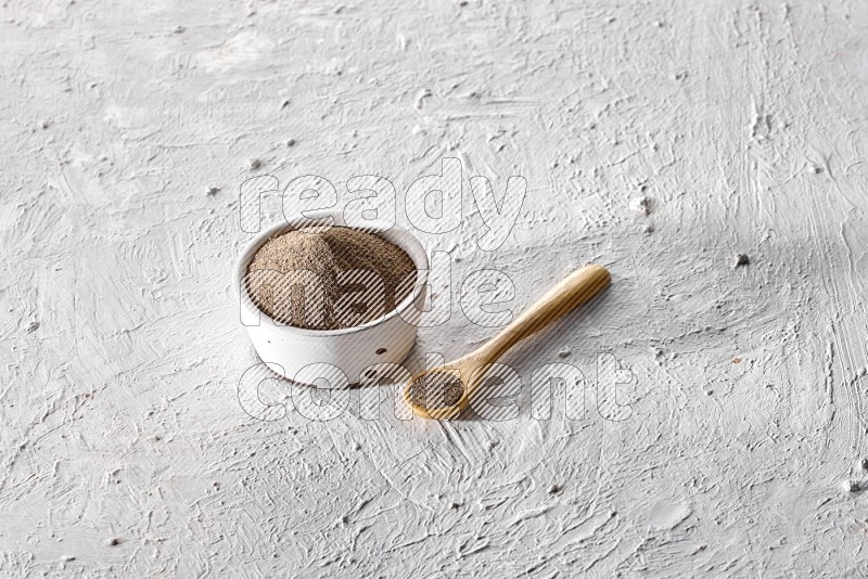 A pottery white bowl full of black pepper powder and wooden spoon full of powder on textured white flooring