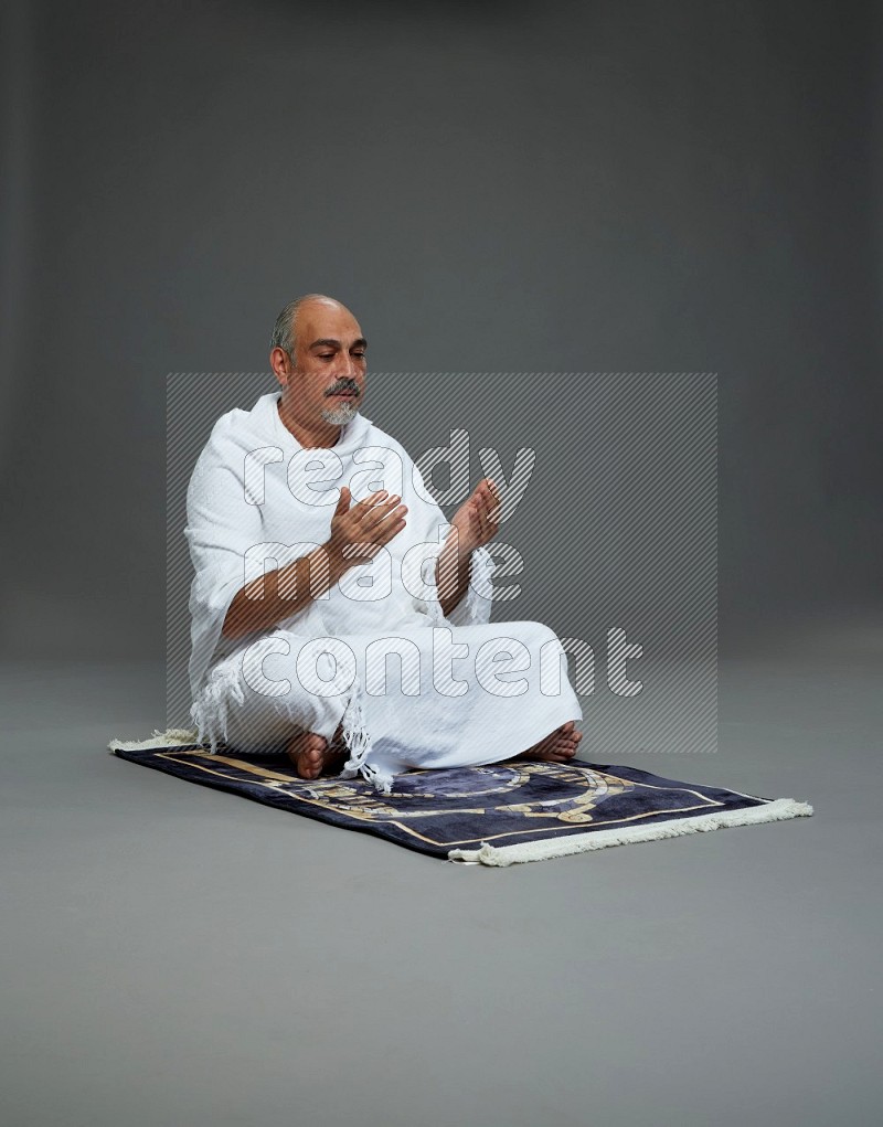 A man wearing Ehram sitting on prayer mat dua'a on gray background