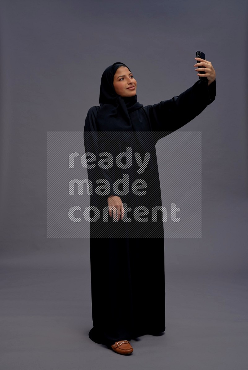 Saudi woman wearing Abaya standing taking selfie on gray background