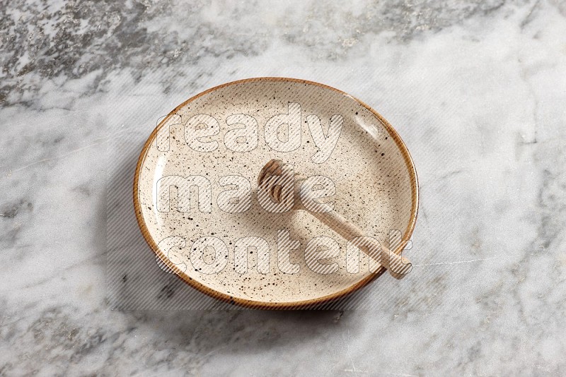Beige Pottery Plate with wooden honey handle in it, on grey marble flooring, 65 degree angle