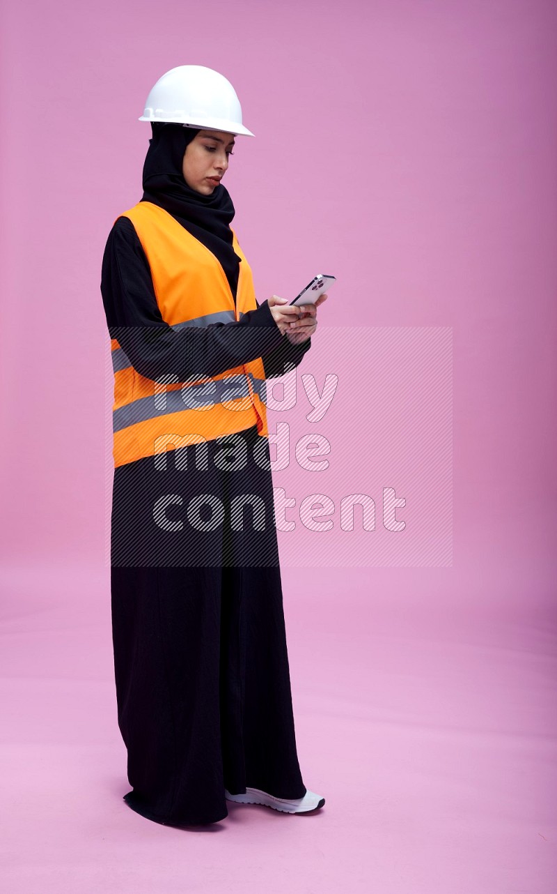 Saudi woman wearing Abaya with engineer vest and helmet standing texting on phone on pink background