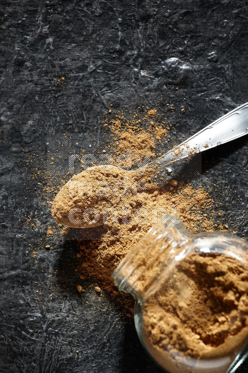 A flipped glass spice jar and a metal spoon full of allspice powder and powder spilled out of it on a textured black flooring