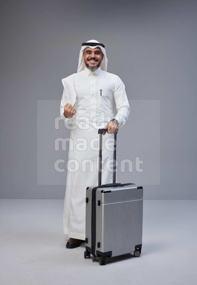 Saudi man wearing Thob and white Shomag standing holding Travel bag on Gray background