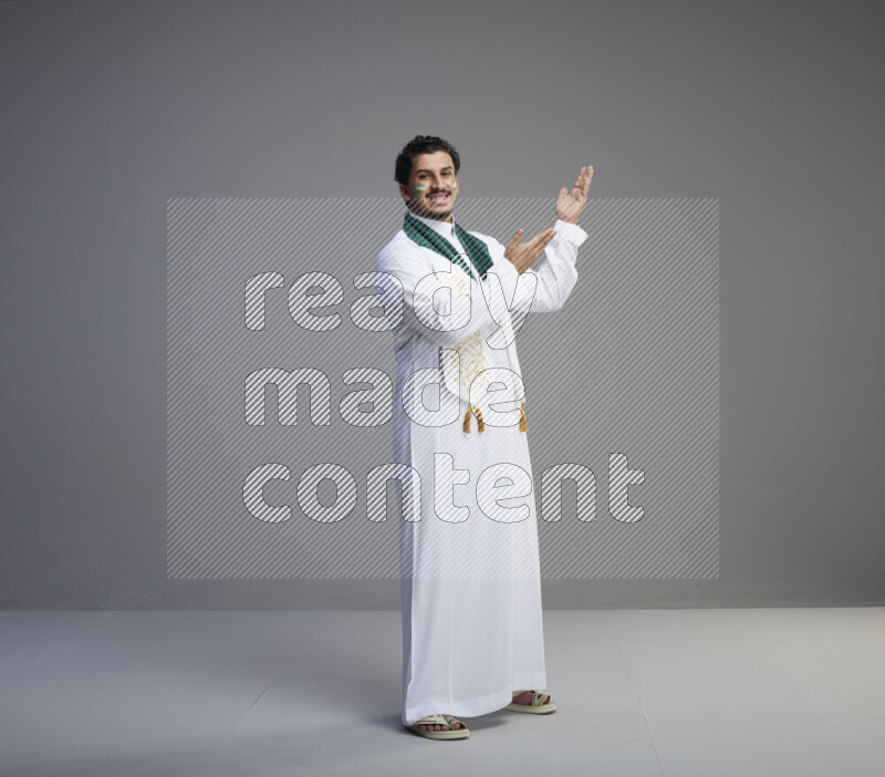 A Saudi man standing wearing thob and Saudi flag scarf with face painting on gray background