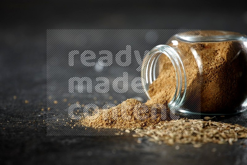 A flipped glass spice jar full of cumin powder with spilled powder and cumin seeds on a textured black flooring
