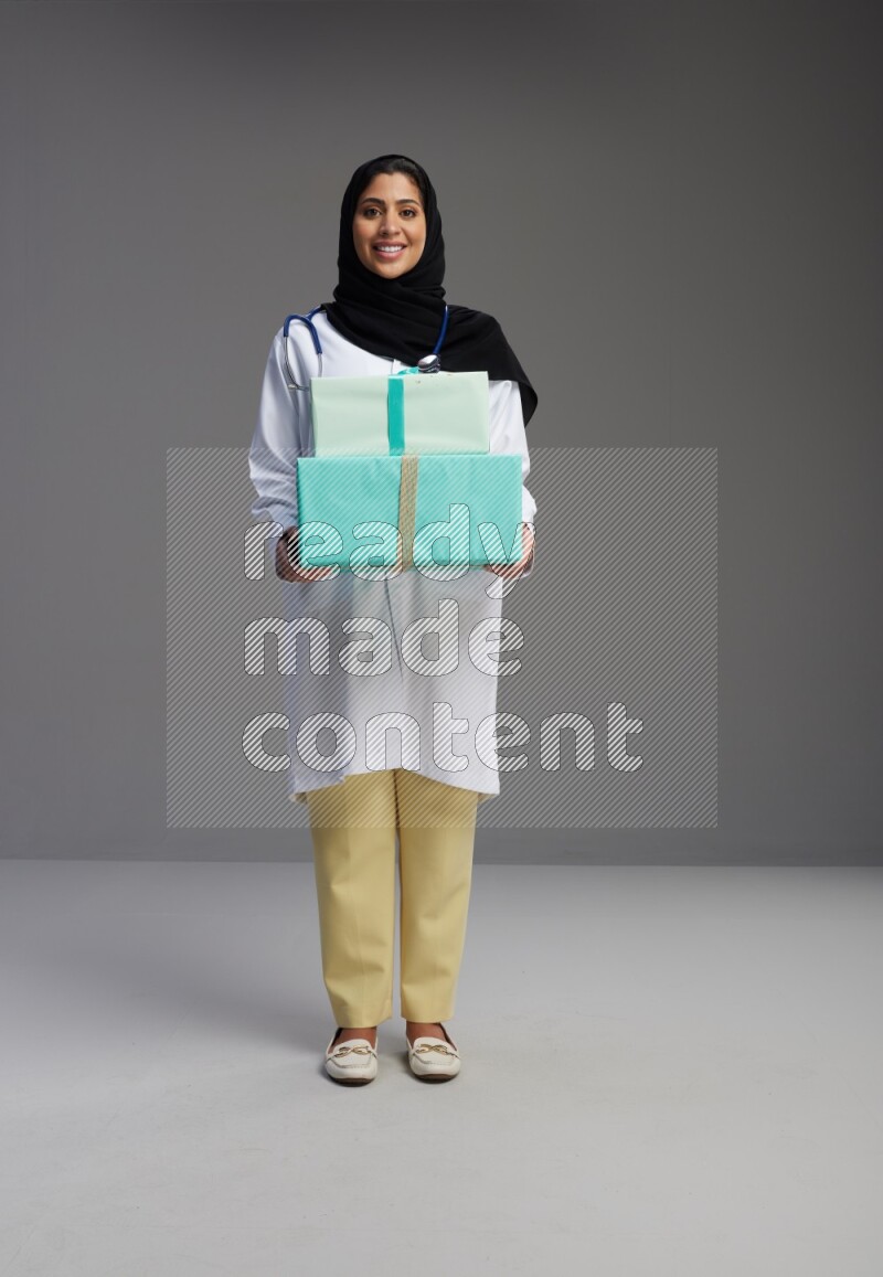 Saudi woman wearing lab coat with stethoscope standing holding gift box on Gray background