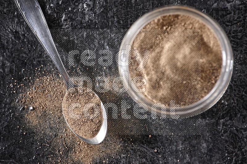 A glass jar full of black pepper powder and a metal spoon full of powder on a textured black flooring
