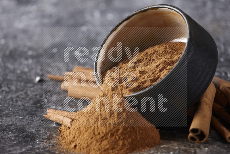Black pottery bowl over filled with cinnamon powder and cinnamon sticks around the bowl on a textured black background