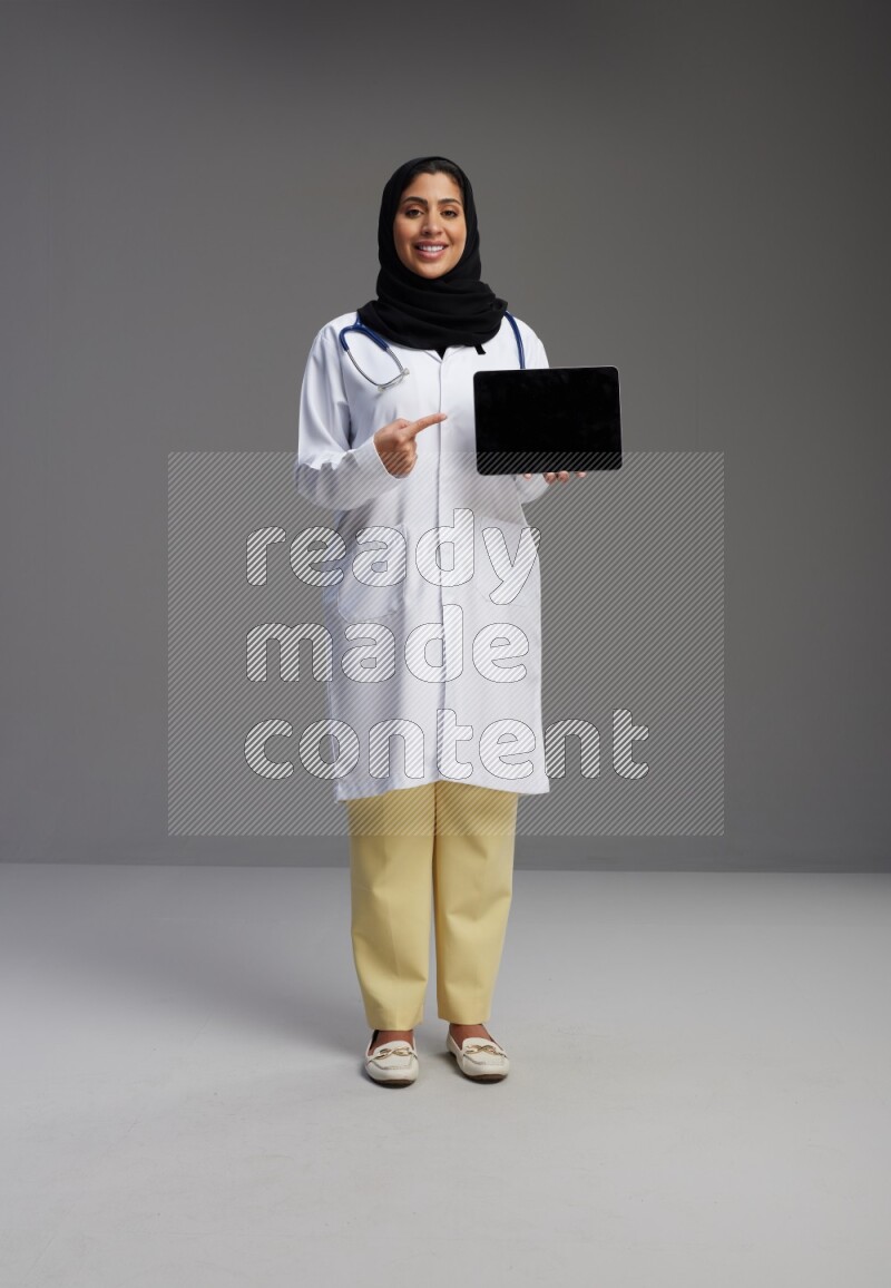 Saudi woman wearing lab coat with stethoscope standing showing tablet to camera with sign in the back on Gray background