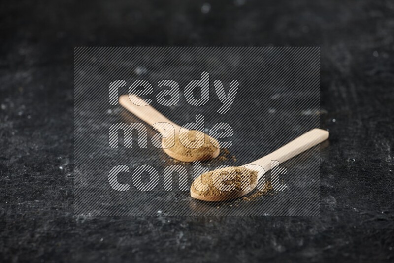 2 wooden spoons full of cumin powder on textured black flooring