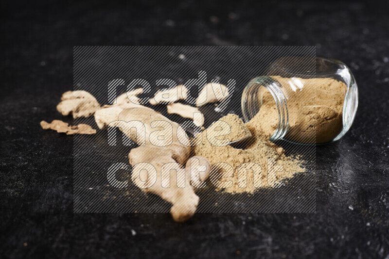 A glass jar full of ground ginger powder flipped with some spilling powder on black background