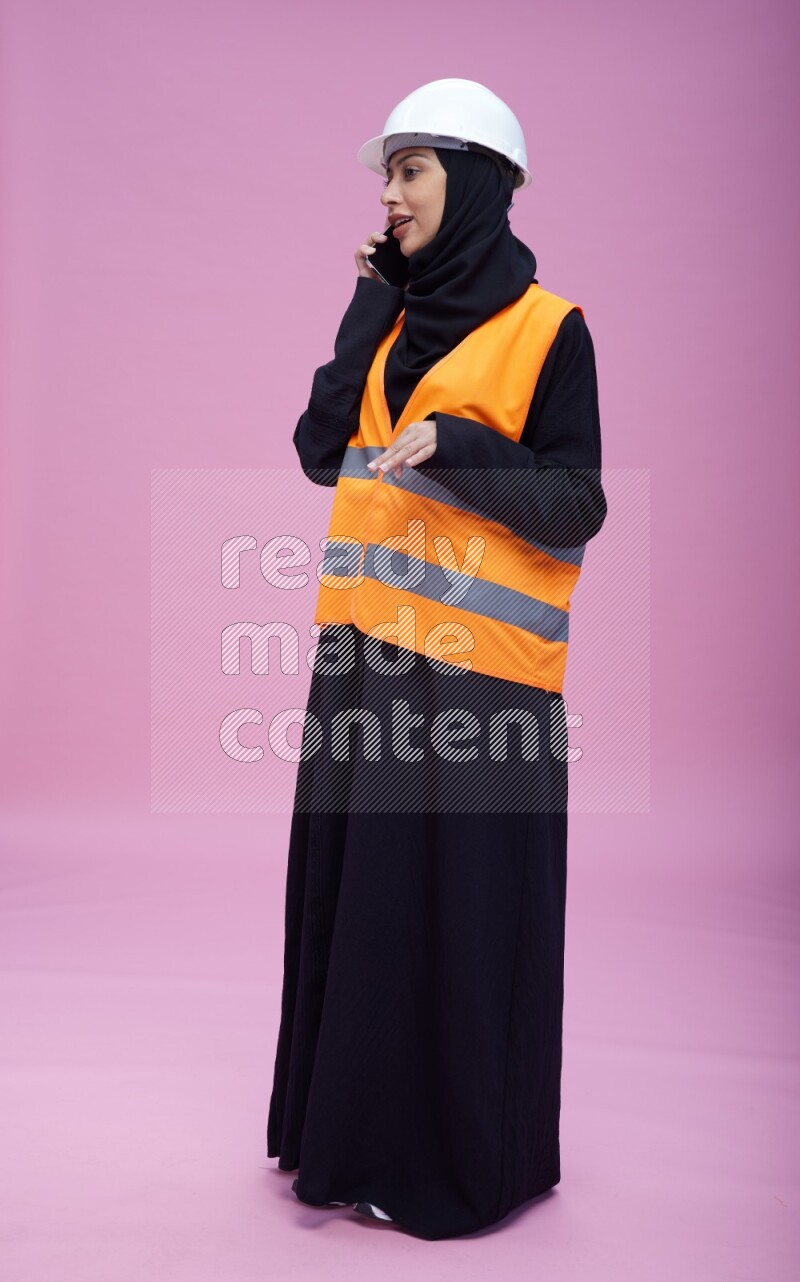 Saudi woman wearing Abaya with engineer vest and helmet standing talking on phone on pink background