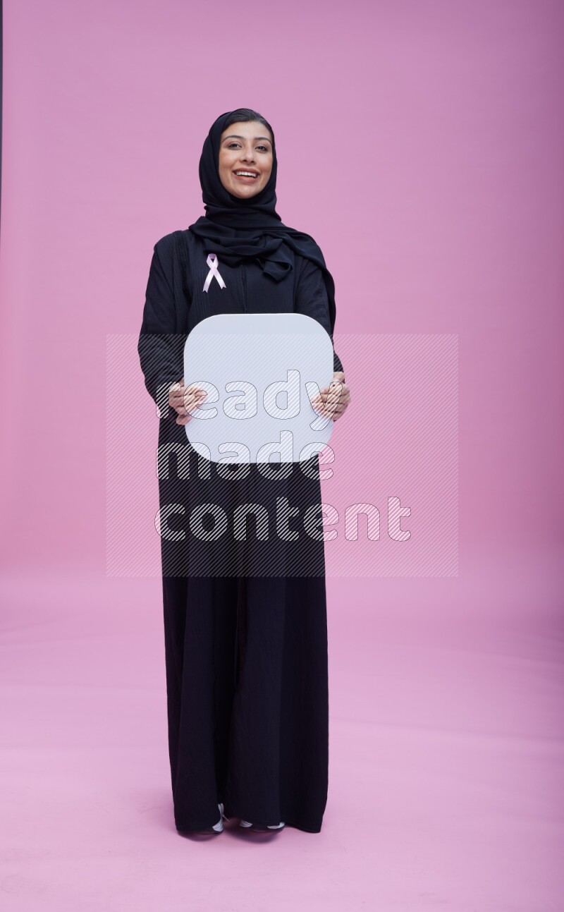 Saudi woman wearing pink ribbon on Abaya standing holding social media signs on pink background