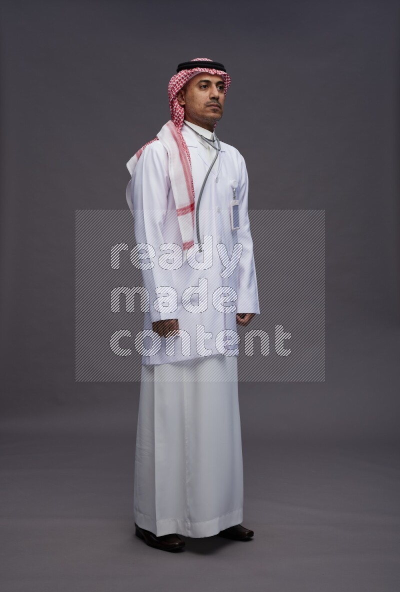 Saudi man wearing thob with lab coat and shomag with pocket employee badge with stethoscope standing interacting with the camera on gray background
