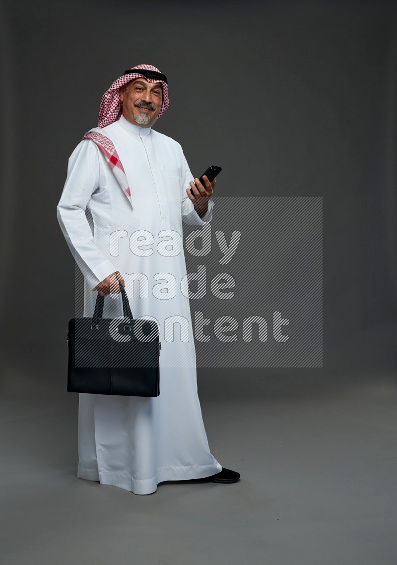 Saudi man with shomag Standing holding bag and phone on gray background