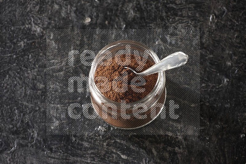 A glass jar full of cloves powder with a metal spoon on a textured black flooring