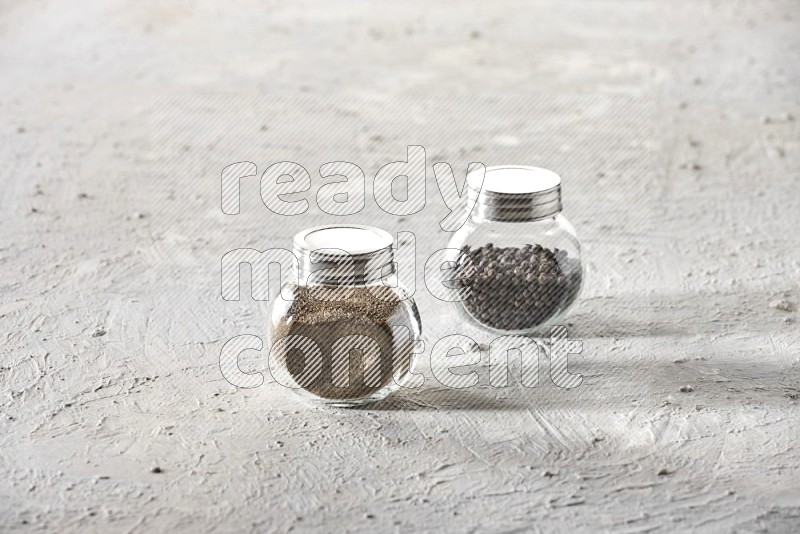 2 glass spice jars full of black pepper and black pepper powder on textured white flooring