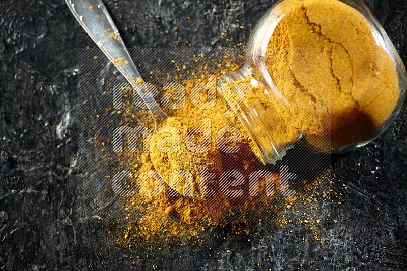 A flipped glass spice jar and a metal spoon full of turmeric powder and powder spilled out of it on textured black flooring