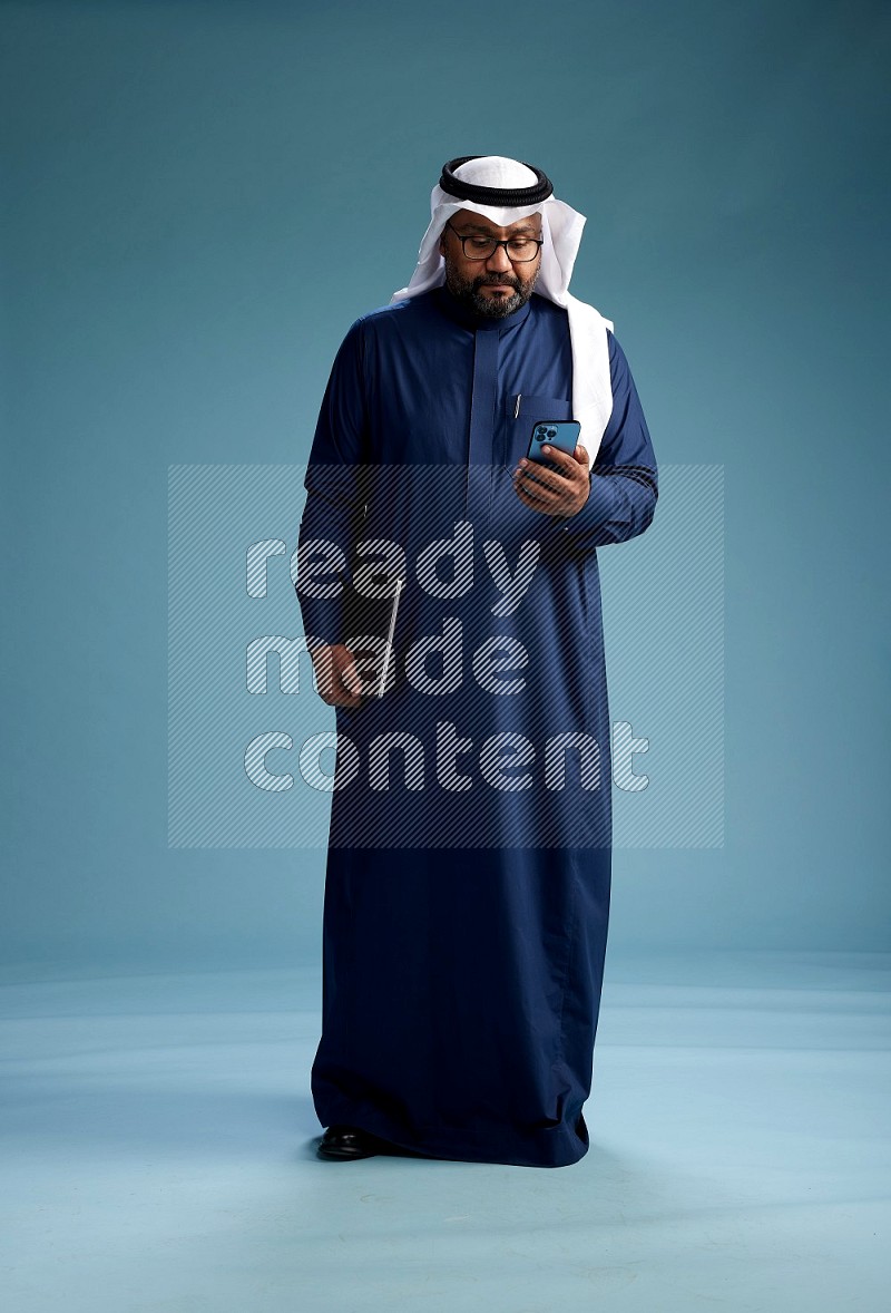 Saudi Man with shimag Standing texting on phone on blue background
