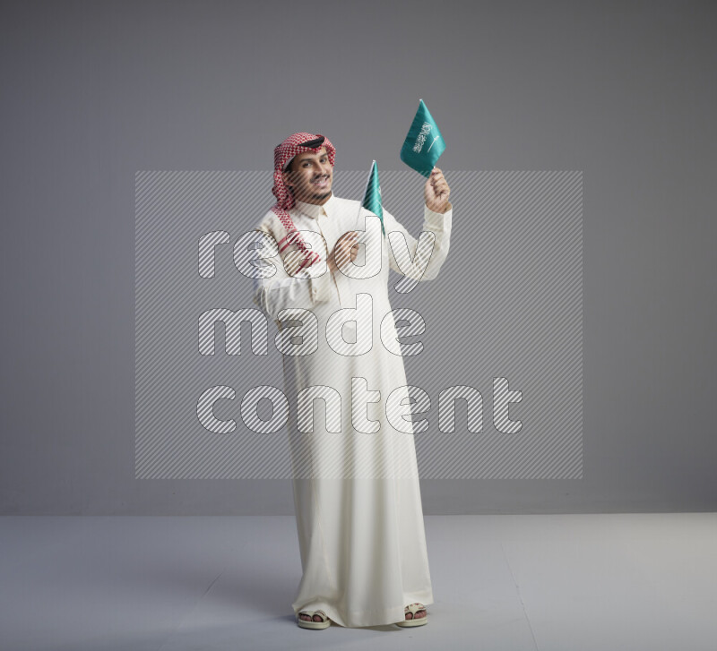 A Saudi man standing wearing thob and red shomag raising small Saudi flag on gray background