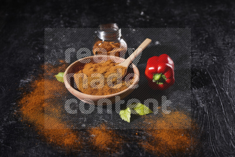 A wooden bowl full of ground paprika powder with a glass jar beside it and a red bell pepper on black background