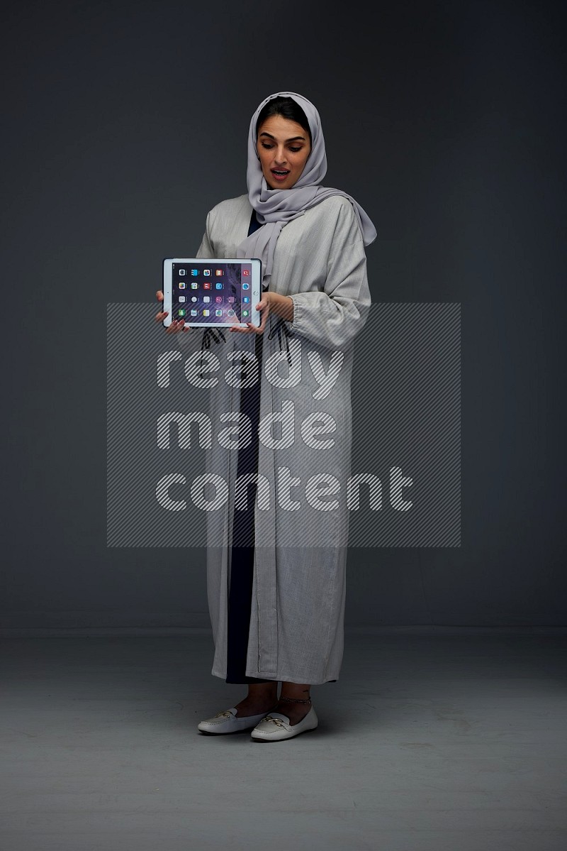 A Saudi woman wearing a light gray Abaya and head scarf standing and showing the phone's screen eye level on a grey background
