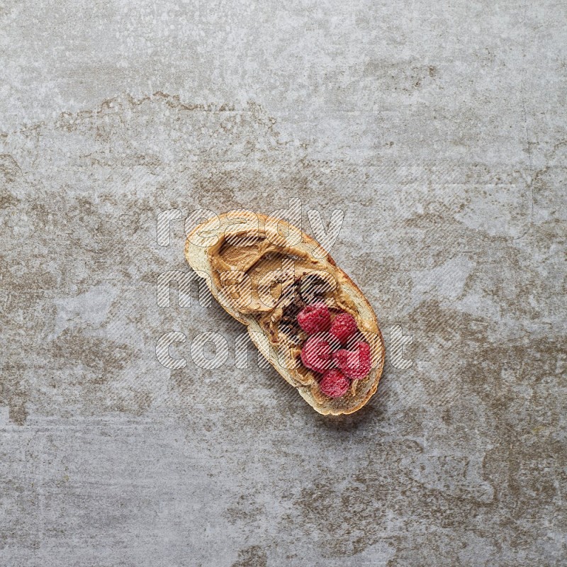 open faced peanut butter sandwich with raspberries and chocolate granula on a grey textured background