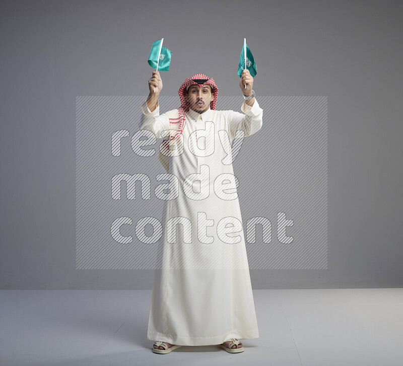 A Saudi man standing wearing thob and red shomag raising small Saudi flag on gray background