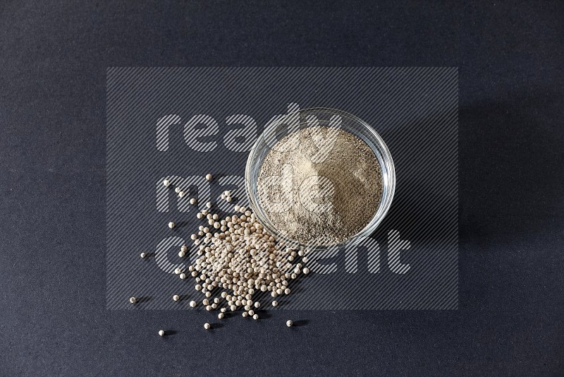 A glass bowl full of white pepper powder with white pepper beads on black flooring