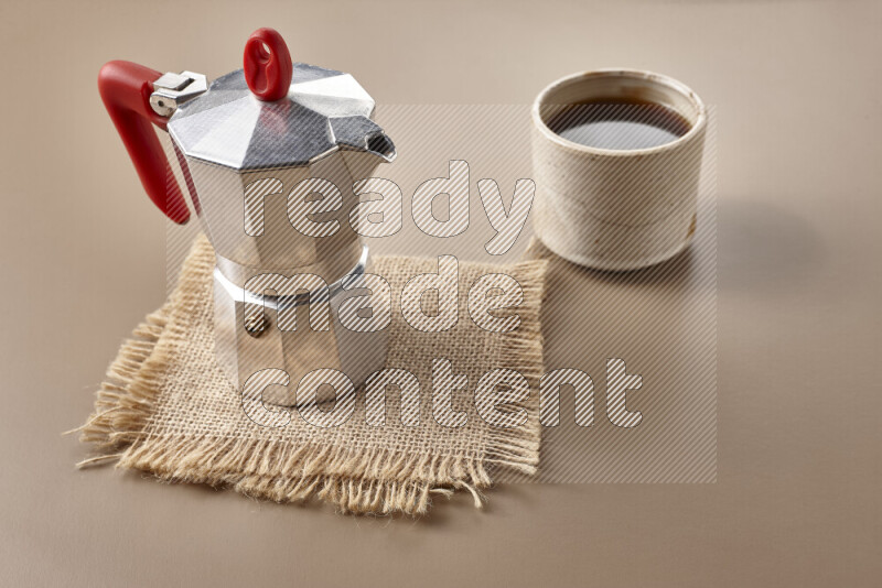 A moka pot with red handle surrounded by roasted coffee beans on beige background