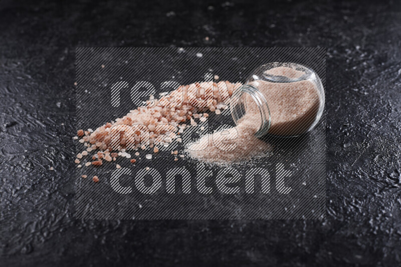 A glass jar full of fine himalayan salt with some himalayan crystals beside it on a black background