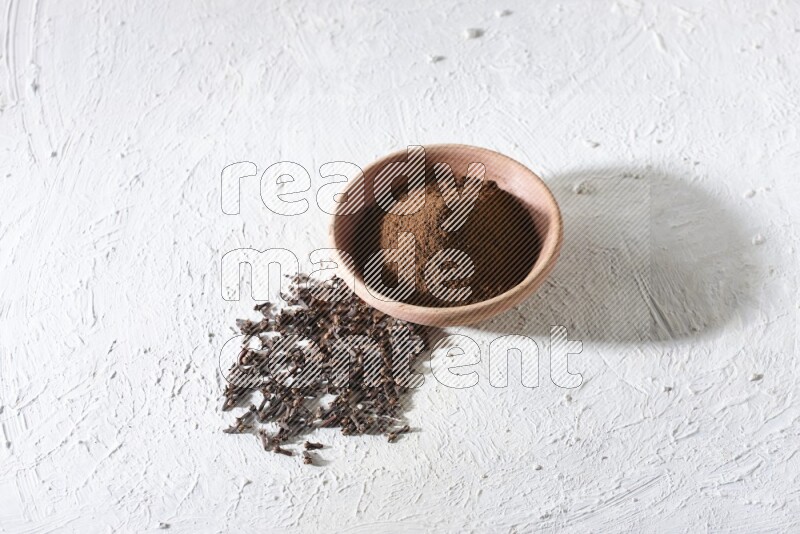A wooden bowl full of cloves powder with whole cloves beside it on a textured white flooring