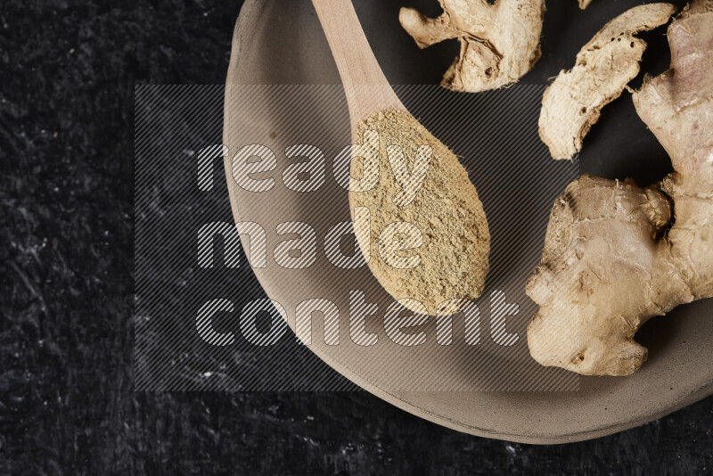 A wooden spoon full of ground ginger powder with fresh and dried ginger, all on a pottery plate on black background