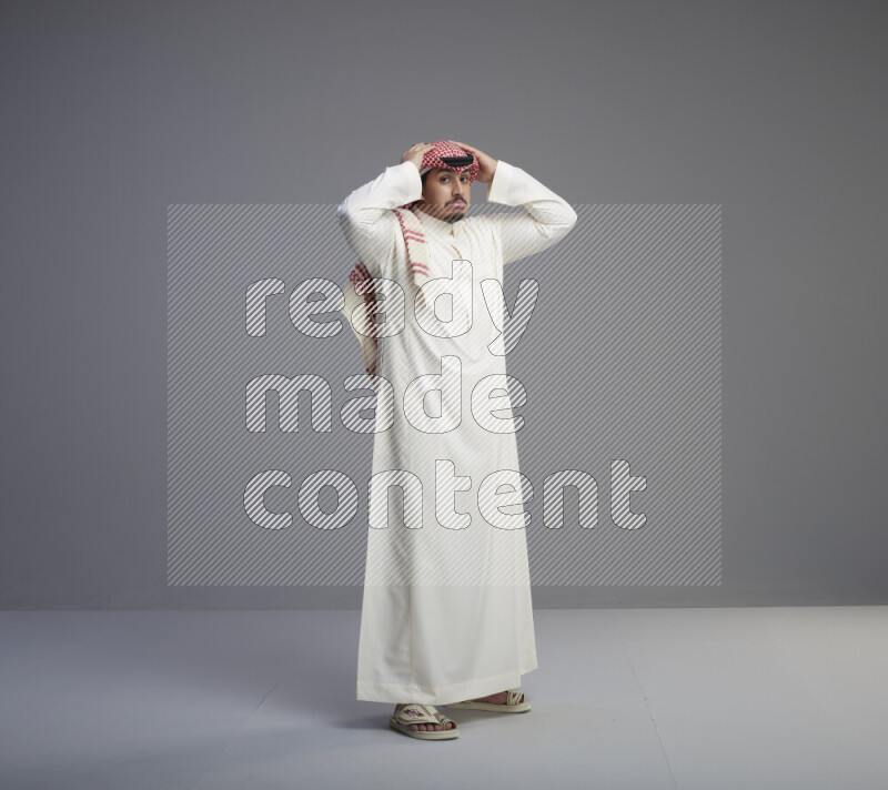 A Saudi man standing wearing thob and red shomag interacting with the camera on gray background