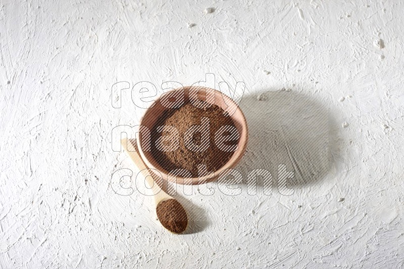 A wooden bowl and a wooden spoon full of cloves powder on a textured white flooring