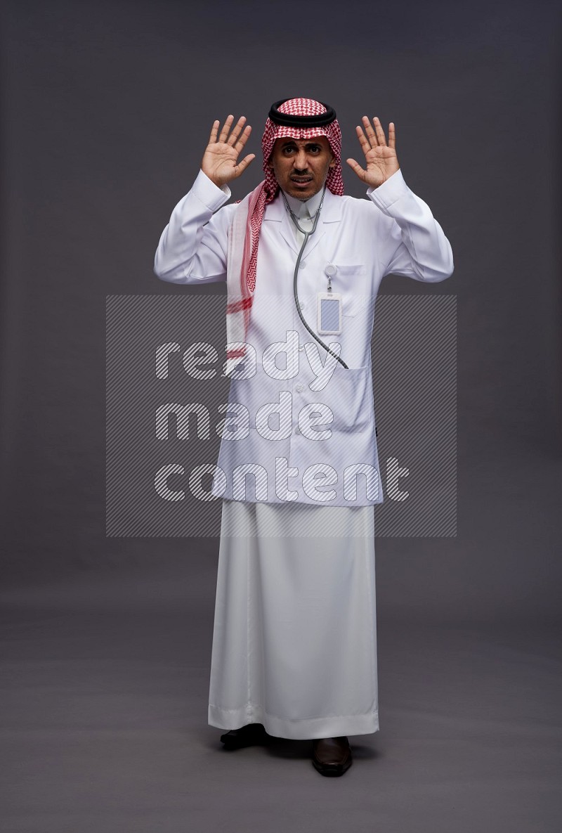 Saudi man wearing thob with lab coat and shomag with pocket employee badge with stethoscope standing interacting with the camera on gray background