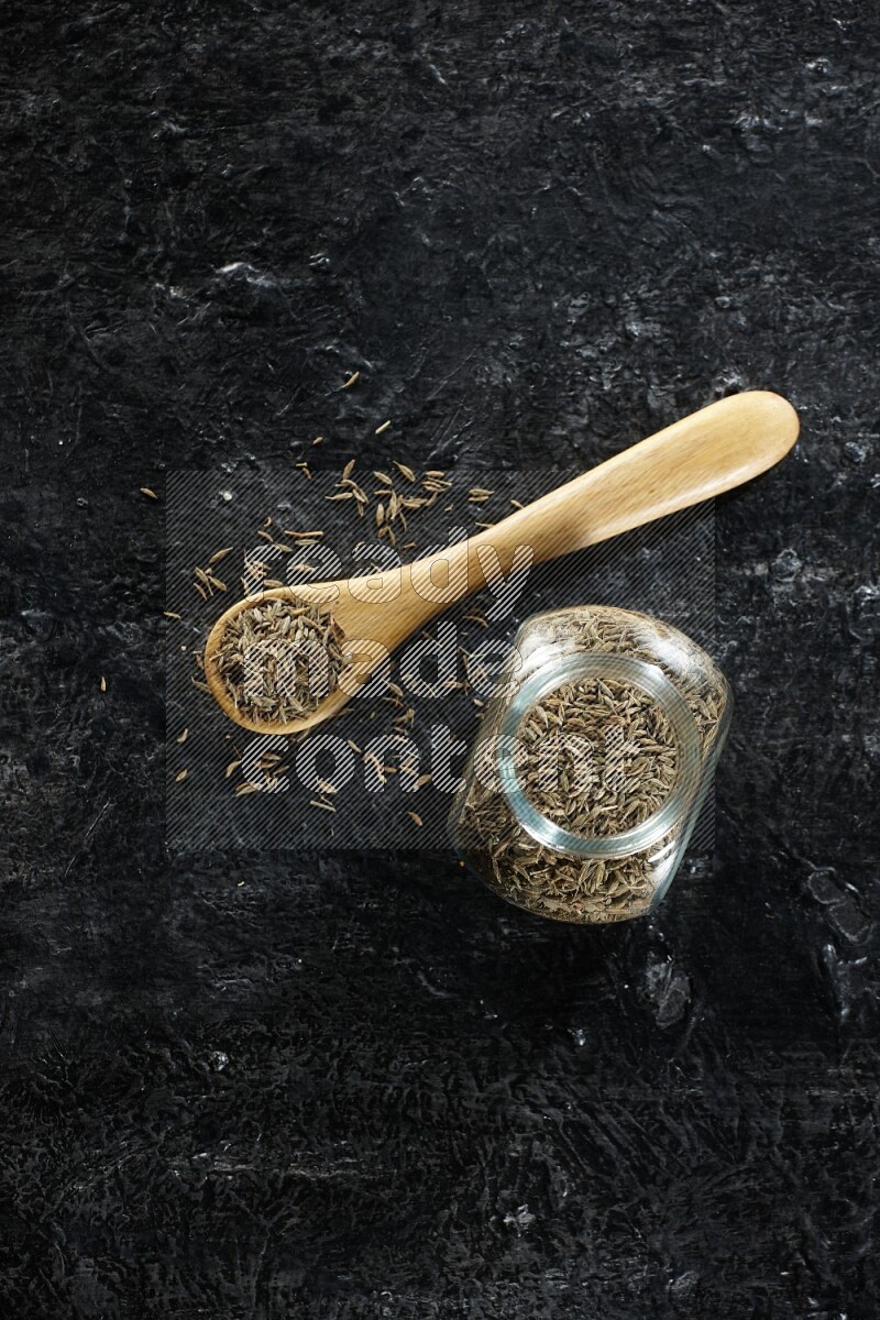 A glass spice jar and a wooden spoon full of cumin seeds on a textured black flooring