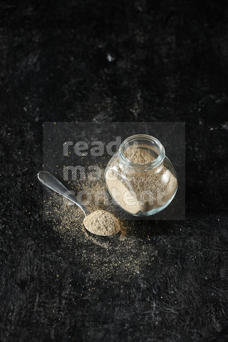 A glass spice jar and metal spoon full of cardamom powder on textured black flooring