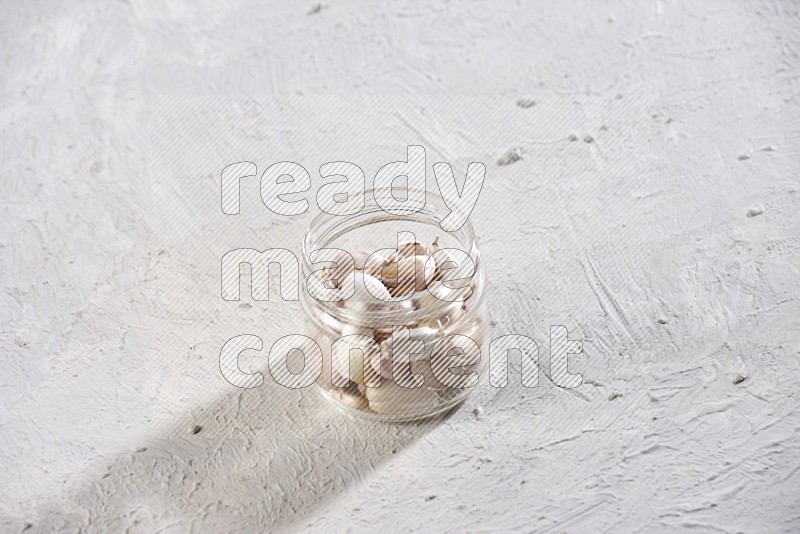 A glass jar full of garlic cloves on a textured white flooring in different angles