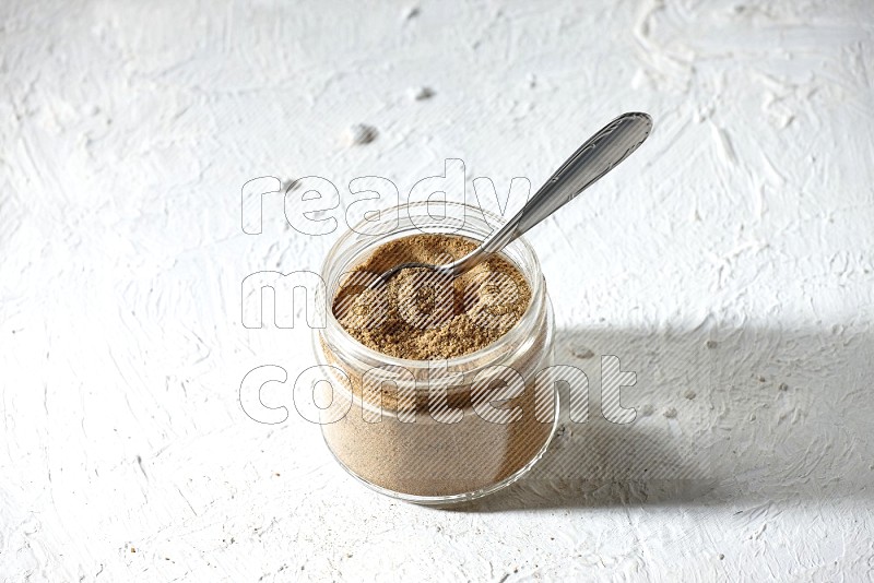 A glass jar and a metal spoon full of cumin powder on textured white flooring