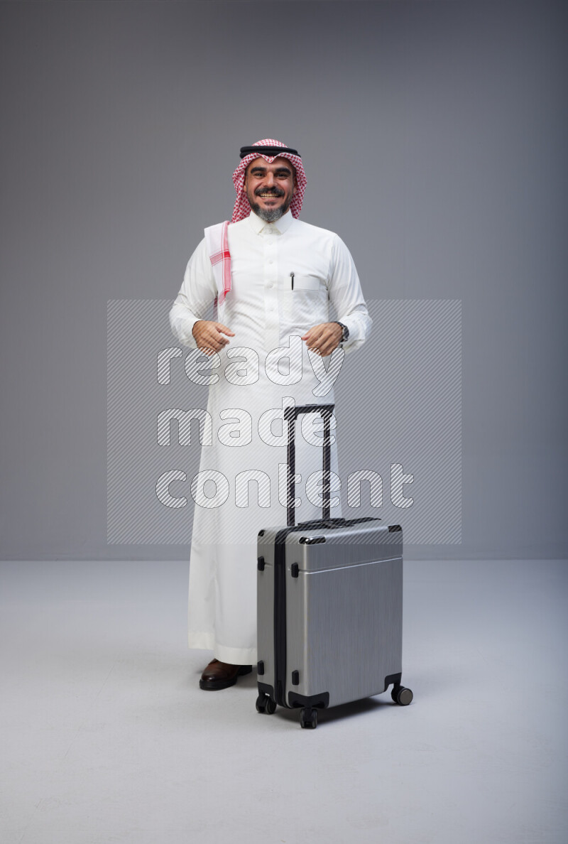 Saudi man wearing Thob and red Shomag standing holding Travel bag on Gray background