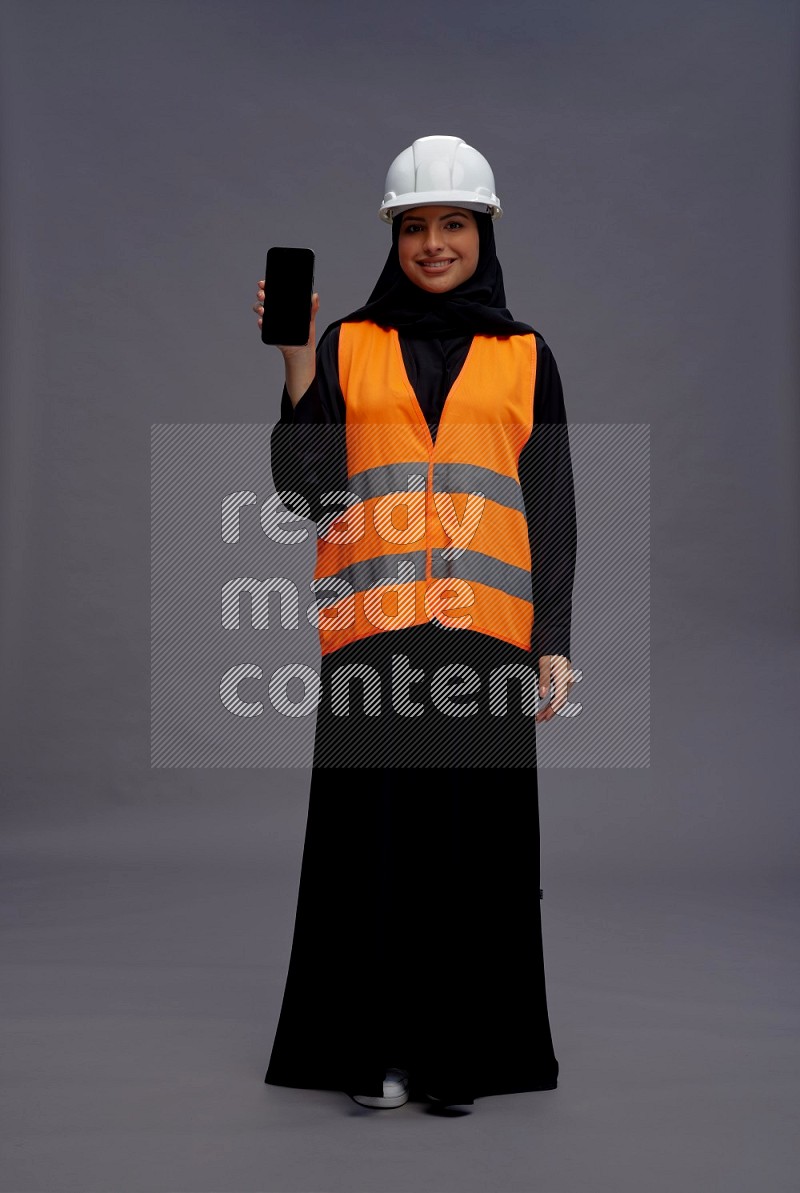 Saudi woman wearing Abaya with engineer vest standing showing phone to camera on gray background